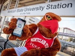 Mascot, Rocky, sits on a bench at the Montclair State University train station. He is showing the NJ Transit app on his mobile phone.