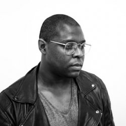 Black and white photo portrait of artist Damien Davis, who wears a black leather jacket, a gray v-neck shirt underneath, and silver framed glasses.