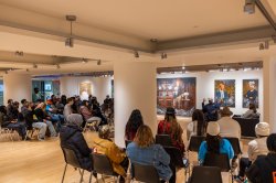 Event photography of an artist talk in the George Segal Gallery. Two speakers on black leather chairs face an audience comprising of students and community members seated on black chairs.