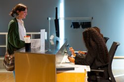 A visitor checks in at the Segal Gallery's front desk with the assistance of two student gallery attendants who sit on office chairs.