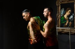 Two dancers wearing shimmery green, gold, and red clothing perform in front of a painting of Angel Gabriel by Carlo Dolci and a work by Joseph Liatela modeled after the railing in the PULSE nightclub. The background is a dark, matte black.