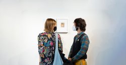 Two visitors wearing face masks look at a work on a white wall.