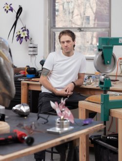 Portrait of artist Justin Cloud seated and wearing a white t-shirt, black pants, and a black wristband. Surrounding him are his artworks and incomplete pieces.