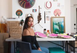 Portrait of artist Caroline Garcia, a woman wearing an off-shoulder long-sleeved black top, blue jeans, red lipstick, and earrings. Behind her is various artworks and materials for creating art.