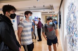 Three students wearing face masks look at a piece of art on the wall.