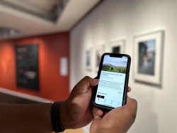 Two hands cradle an iPhone with the University Galleries' digital guide loaded on the screen. In the background, on the right, are framed photos on a white wall, and on the left, a black and red wall with fake weapons.
