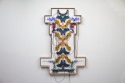 An artwork hangs on a white wall. The work is made up of circuit boards of various colors, including teal, mustard yellow, and plum red. The work has silver hardware and black wires that cross over one another.