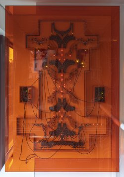 An artwork made of circuit boards and wires behind orange acrylic is affixed to the wall.