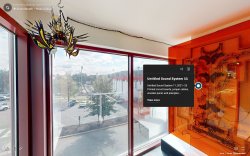 Screenshot of a virtual walkthrough of the Sound Booth installation. On the left is an artwork made of circuit boards and wires behind orange acrylic is affixed to the wall. On the right hangs a small, black and yellow artwork with orange-yellow lights and sharp edges.