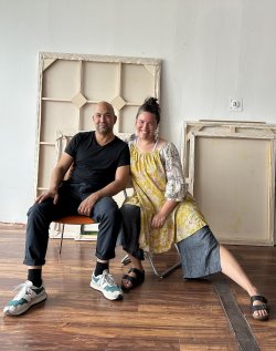 Portrait of a man, left, and woman, right, both seated and smiling. Behind them are blank canvases of varying size.