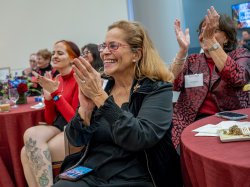 A woman in a black shirt with blonde hair and glasses smiles widely while clapping