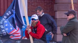 Photo of man holding a QAnon flag, wearing red sweatshirt, sunglasses and hat. Two men stand in the background