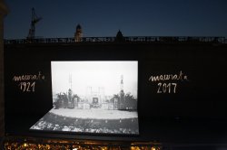 Wide shot of a theater stage with a black and white image projected. Hand written text on the screen says "Macerata 1921" and "Macerata 2017."