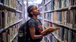 student standing between two book shelves in library, holding book and looking up at shelf