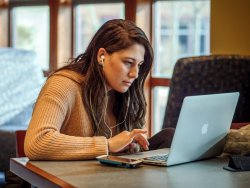 photo of white college aged woman working on laptop with headphones