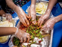 several pairs of hands wearing gloves rub spices onto green onions in an aluminum pan