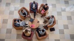 Seven graduate students collaborating while seated in a circle with laptops and papers