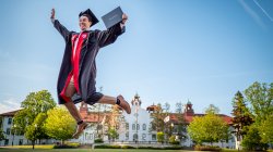graduate in cap-and-gown leaping through the air with diploma in hand