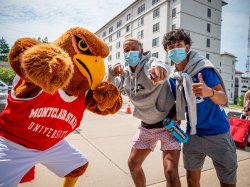 Two Montclair State students pose with Rocky the Red Hawk in front of a residence hall