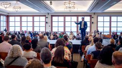 wide view of Dr. Koppell speaking on a raised platform surrounded by faculty in the conference center
