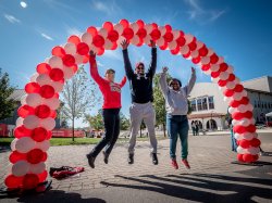 Three alumni jump under a red and white balloon arch