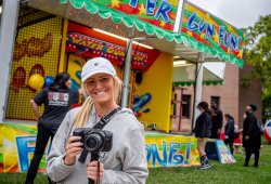 Student photographer pauses with camera on monopod in front of carnival games