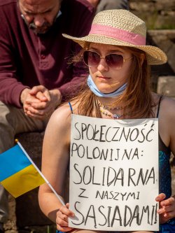 Woman in hat and sunglasses reading a sign in polish reading "społeczność polonijna: solidarna z naszymi sąsiadami"
