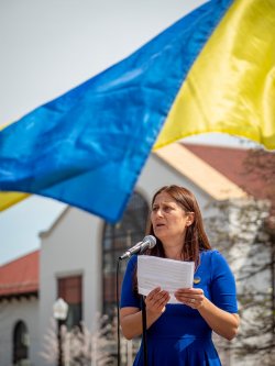 Woman speaking into micrphone with ukranian flag blowing in the breeze above her head