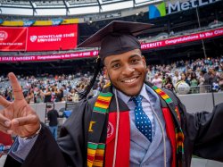 photo of a smiling graduate in a stadium with Montclair State signs behind him