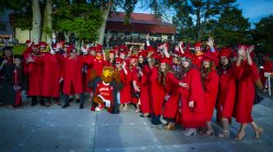 photo of a large group of graduates in red gowns and mortar boards posing with Rocky the Red Hawk mascot