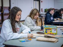 Two female students sit at a table and draw and color on paper bags.