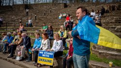 man wearing Ukrainian flag around his shoulders stands near people seated in amphitheater