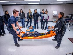 Two student EMT volunteers carry a woman on an orange stretcher while several students watch.