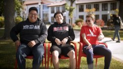 Three young men sit in red chairs on campus.
