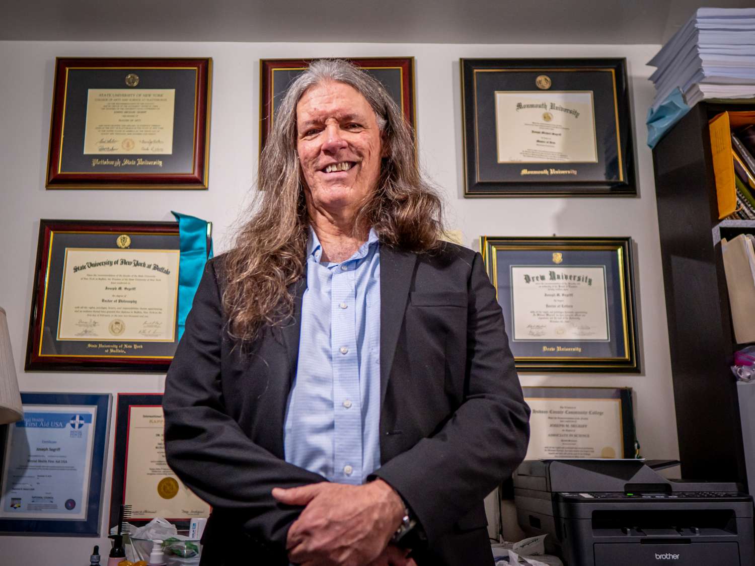 A man stands in front of a wall covered with diplomas.