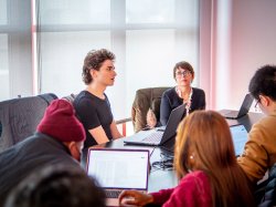 Students sit around a table while a professor looks on in the background.