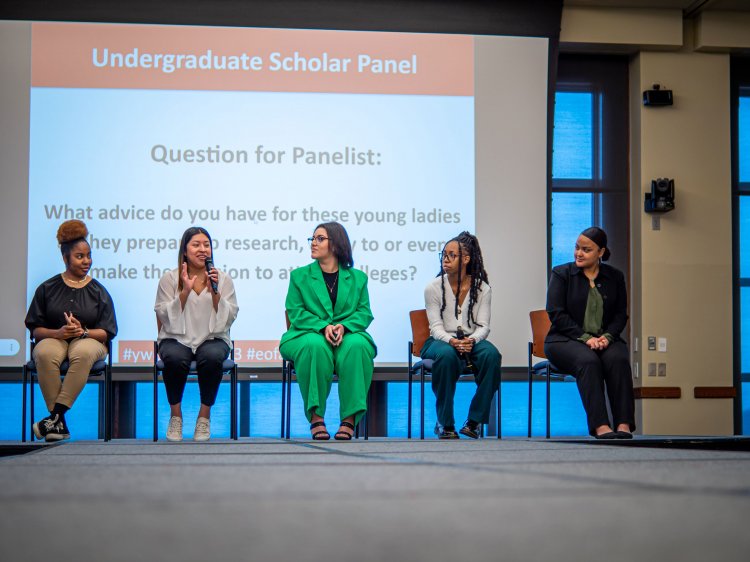 Five female panelists sit on a stage as one of them speaks.
