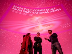 Three people stand in front of a pink screen that reads “Space Talk: Cosmic Clues from Gravitational Waves”