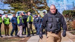 Foreground: police officer in bulletproof vest walking toward camera. Background: a group of police officers in bulletproof vests and reflective vests having a conversation.