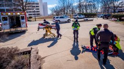 Two paramedics load a man on a stretcher into an ambulance while three more tend to a man on the ground while a police officer stands to the side.