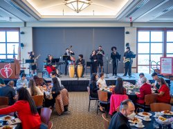 A band with bongos, brass and string instruments performs in front of a large audience at a luncheon