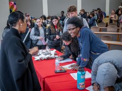 A student smiles and leans forward to talk with a woman, as other students wait their turn in line.