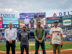 Four men stand in a baseball stadium and also appear on the big screen behind them.