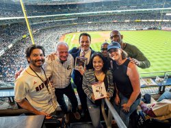 Seven people smile from a baseball box seat with the field below.