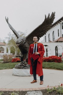 A male student wearing a graduation gown and holding a red mortarboard poses before a large, bronze Red Hawk statue.