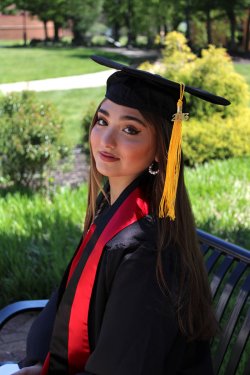 Graduate wearing a graduation cap and gown poses on a bench.