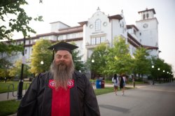 A bearded graduate wearing a cap and gown smiles.