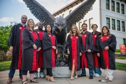 Eight students in graduation gowns pose in front of a bronzed Red Hawk statue.