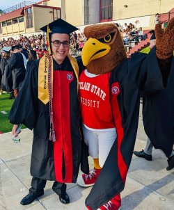 A male student in cap and gown poses with a Red Hawk mascot.