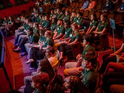 Students in green shirts sit in an auditorium looking toward a stage.